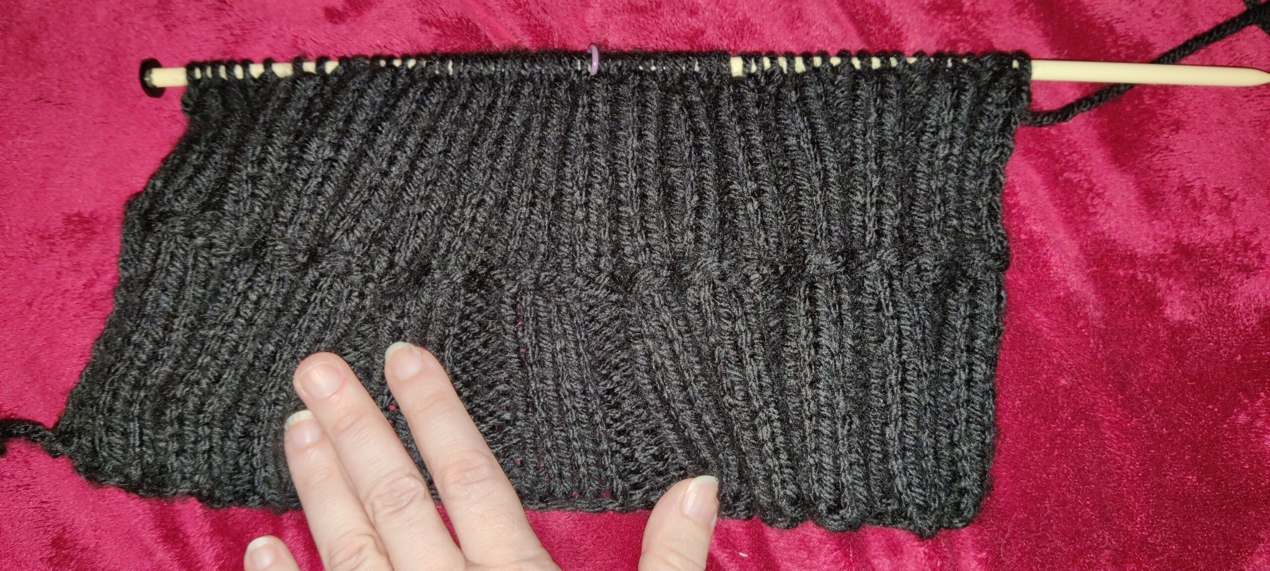 An image of a black piece of knitting in progress, still on the needle. There is a person's hand on top of the piece, so from scale we can tell it is about 5" long with a 2x2 rib and lying on a red background.